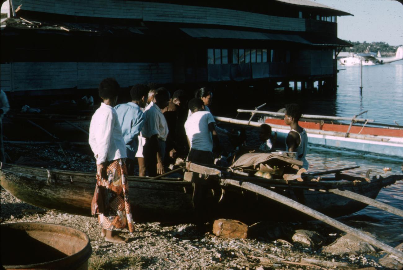 BD/288/95 -
Groep papoea's bij prauw aan de haven met op de achtergrond een watervliegtuig