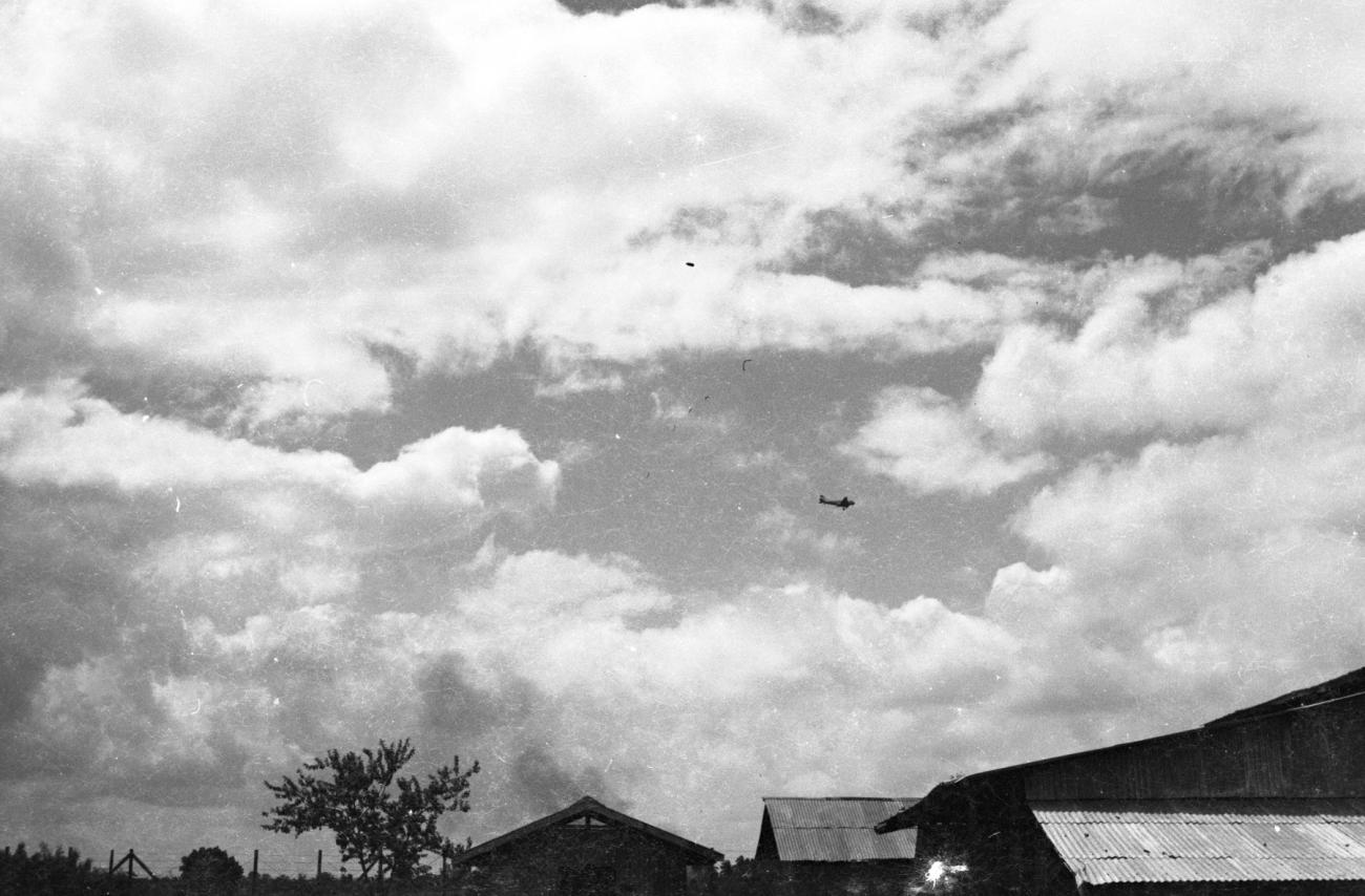 BD/133/1108 -
Overkomend vliegtuig boven golfplaten daken