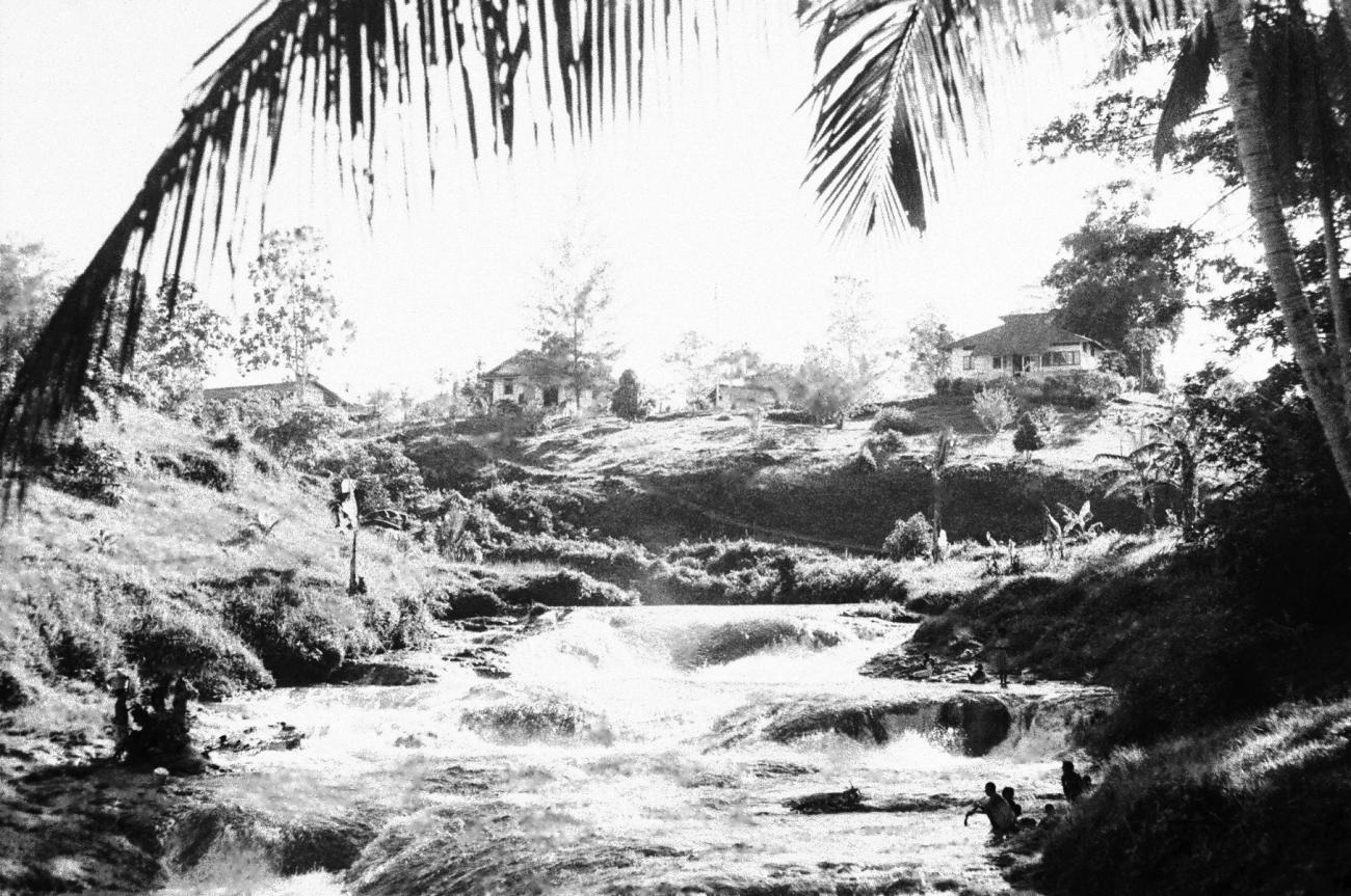 BD/133/679 -
Stroomversnelling in de rivier met nederzetting op de hoge oever en Papoea's langs het water
