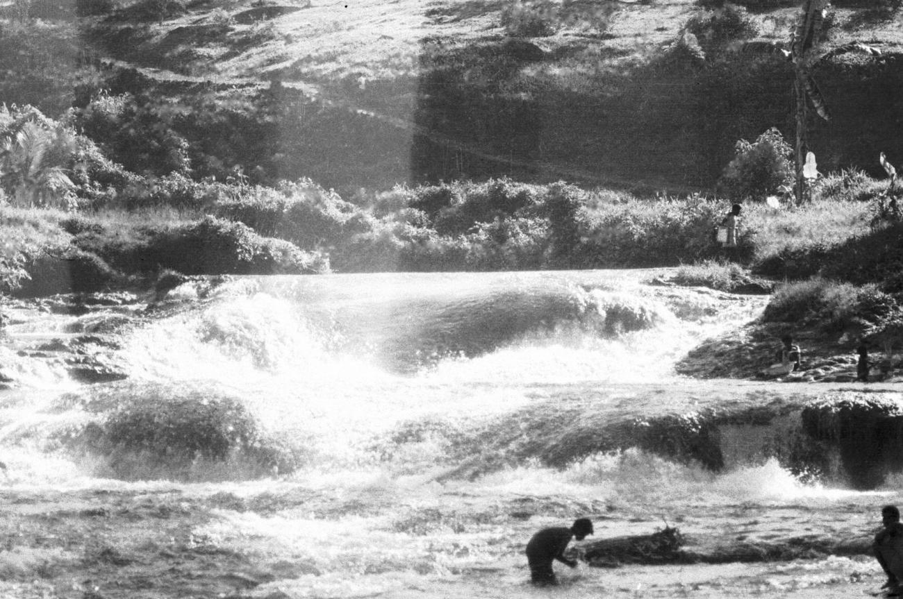 BD/133/680 -
Stroomversnelling met hoge rivieroever en Papoea-vrouwen en kinderen bezig langs het water