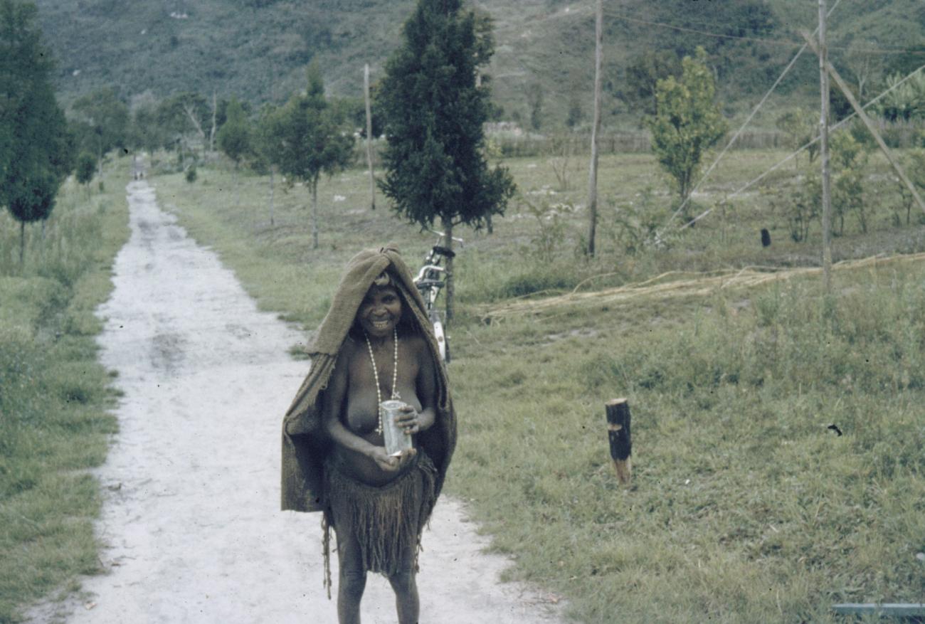 BD/285/84 -
Vrouw met flesje in haar hand en doek om hoofd en ketting om hals poseert op landweg