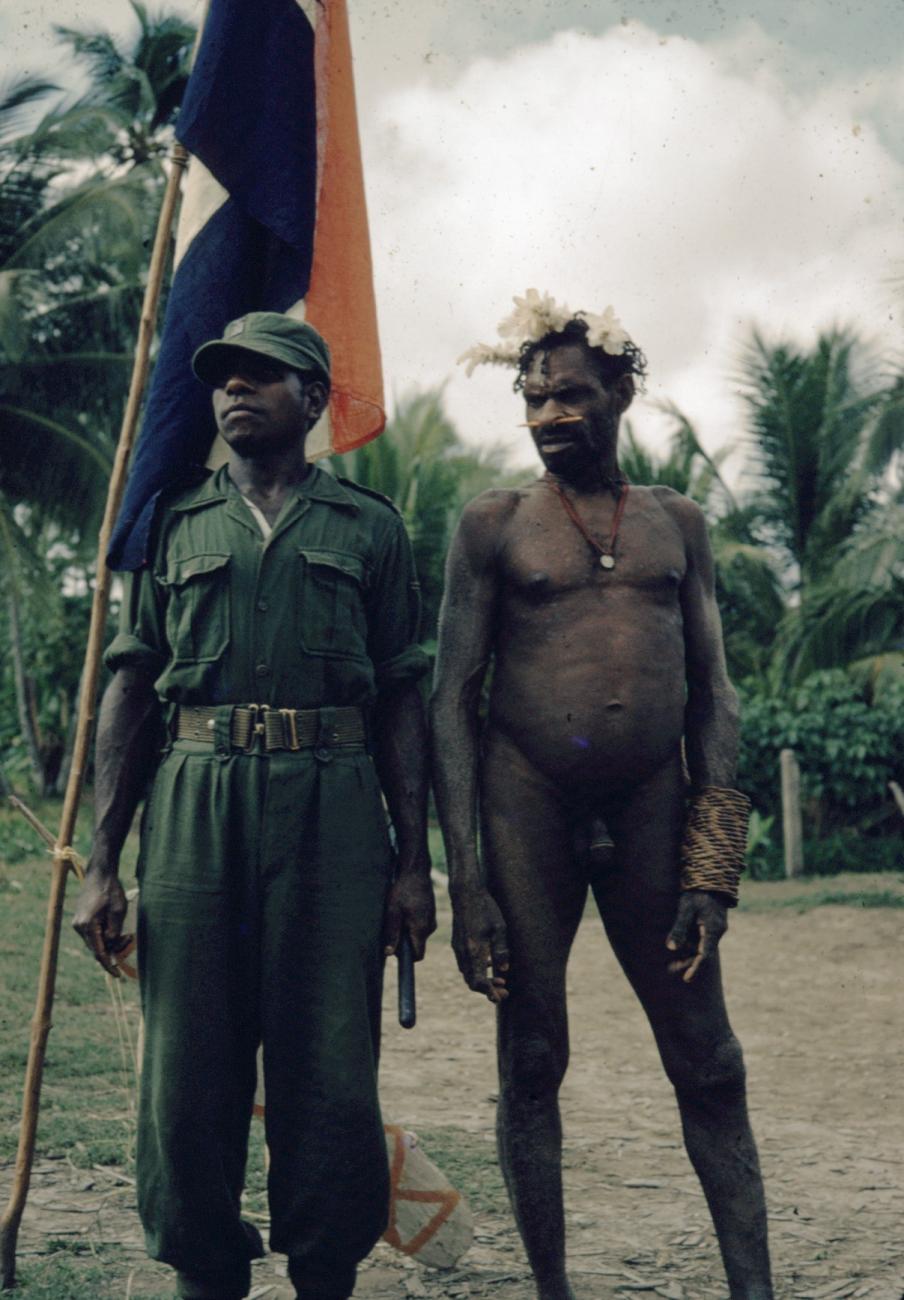 BD/66/135 - 
Asmatter en militair staan naast Nederlandse vlag
