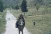 BD/285/84 Vrouw met flesje in haar hand en doek om hoofd en ketting om hals poseert op landweg